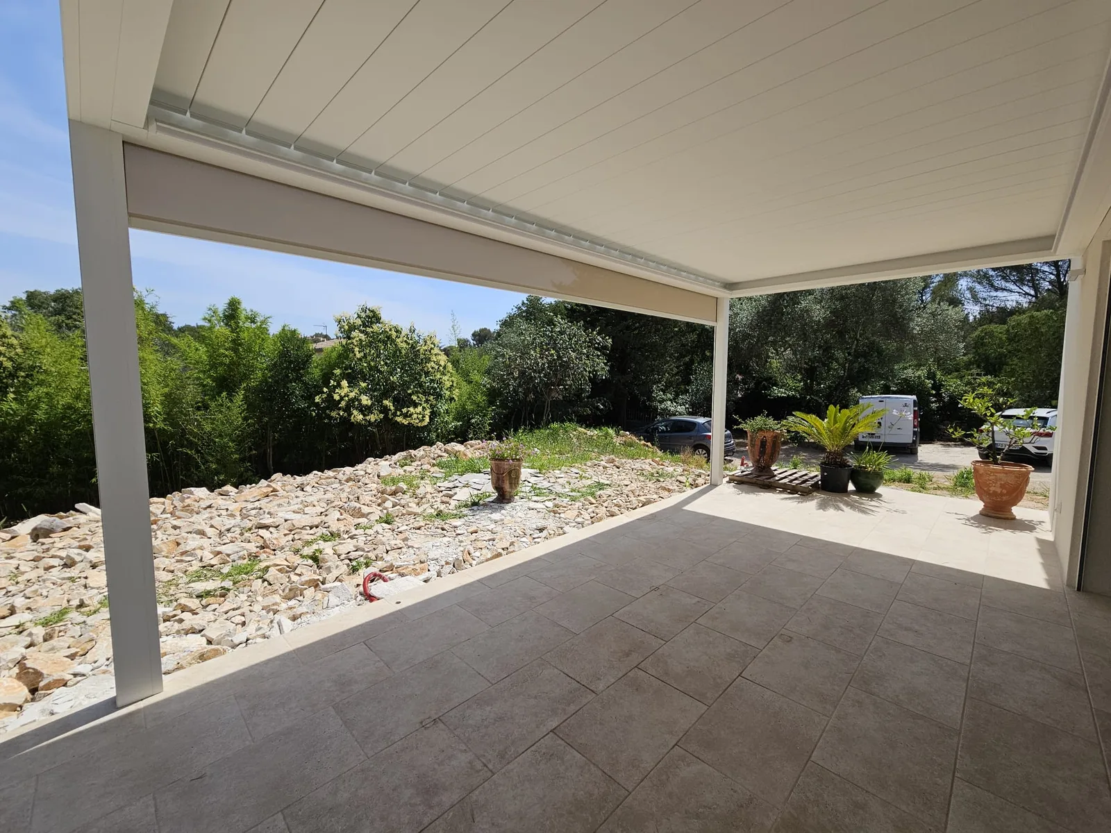 Intérieur de la pergola Brustor avec terrasse carrelée, lames ouvertes et environnement extérieur verdoyant.